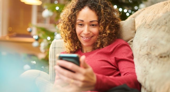 mensagem-de-natal-para-empresas-no-celular Mulher lendo uma mensagem no celular em clima de Natal, representando uma mensagem de natal para empresas enviada de forma humanizada.