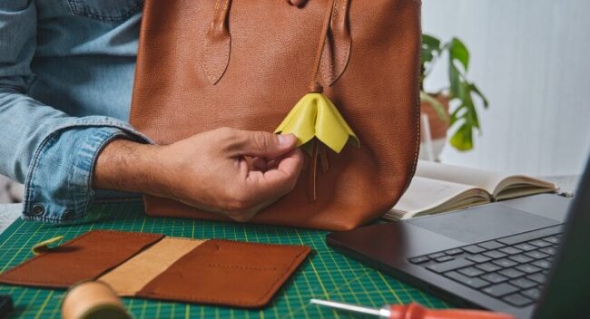 Detalhe de bolsa de couro sendo finalizada à mão em bancada de trabalho, com ferramentas de costura e notebook ao fundo.