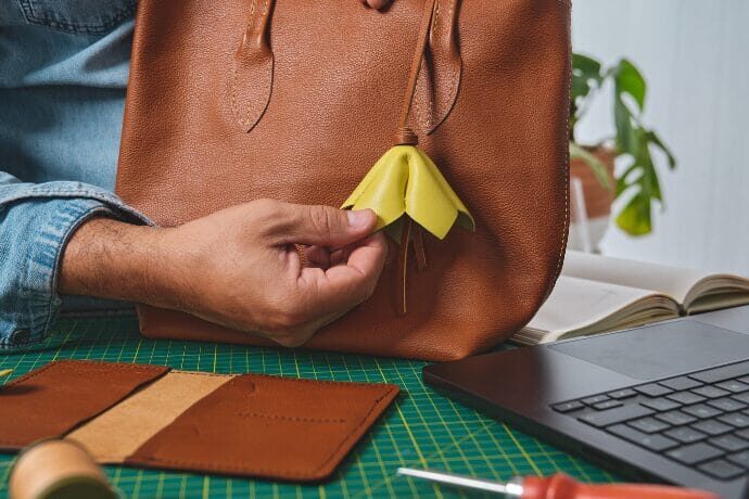 Detalhe de bolsa de couro sendo finalizada à mão em bancada de trabalho, com ferramentas de costura e notebook ao fundo.