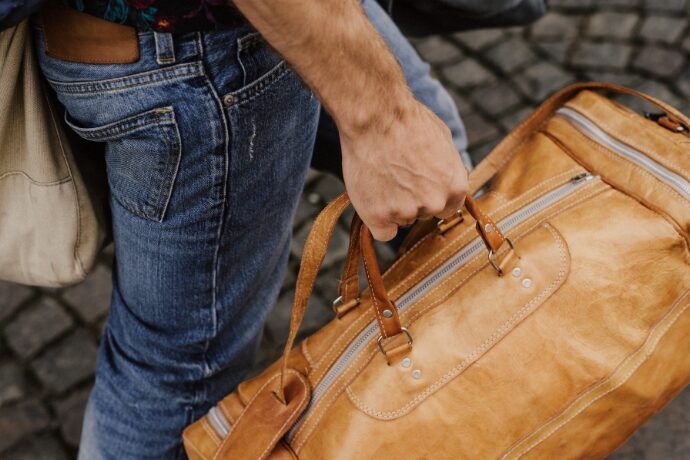 Bolsa de viagem de couro em tom caramelo sendo carregada por homem em ambiente externo, representando estilo de vida sofisticado e deslocamento para o campo.