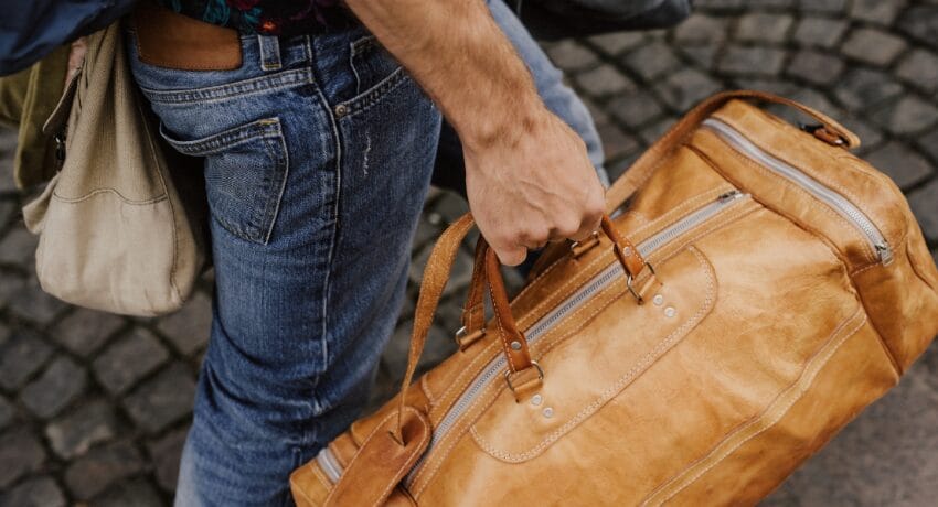 Bolsa de couro para viagem no campo com estética de luxo silencioso Bolsa de viagem de couro em tom caramelo sendo carregada por homem em ambiente externo, representando estilo de vida sofisticado e deslocamento para o campo.