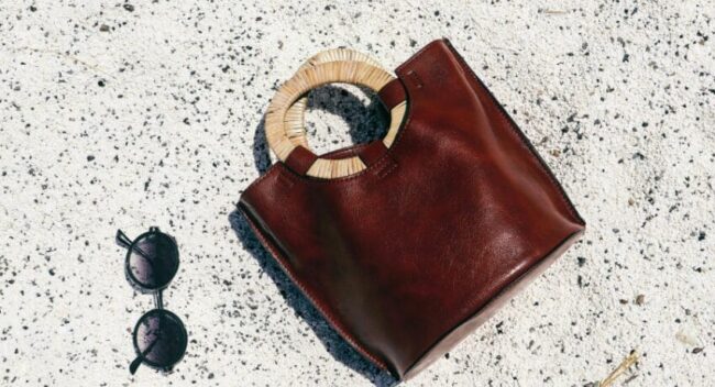 Bolsa de couro sofisticada e acessórios que refletem estilo de vida de luxo Bolsa de couro marrom com alça circular em material natural apoiada sobre piso claro, acompanhada de óculos escuros, representando estilo sofisticado e lifestyle de luxo.