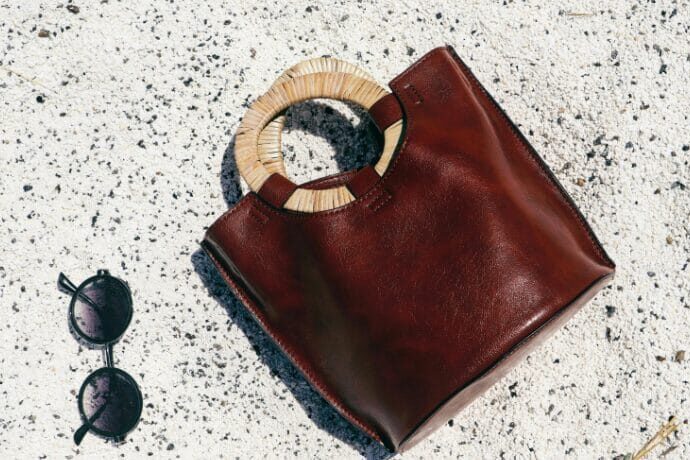 Bolsa de couro marrom com alça circular em material natural apoiada sobre piso claro, acompanhada de óculos escuros, representando estilo sofisticado e lifestyle de luxo.