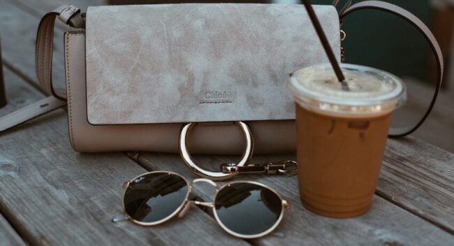 Bolsa feminina de luxo em tom neutro sobre mesa de madeira, acompanhada de óculos de sol e café gelado, em ambiente externo aconchegante.